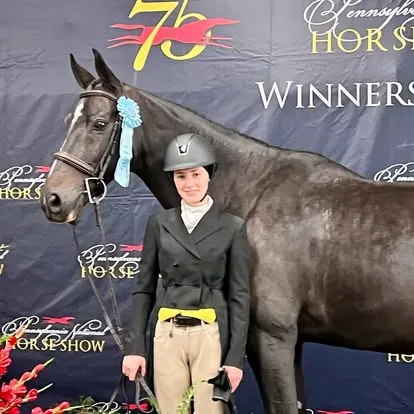 Rider in dark coat beside dark horse with blue ribbon