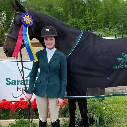 Rider standing with dark horse wearing blue tricolor Saratoga ribbon