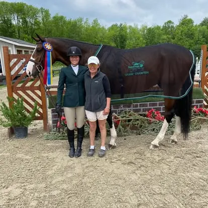 Two women beside blanketed bay horse with tricolor grand champion ribbon