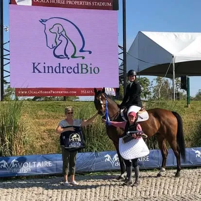Woman leads bay horse below KindredBio jumbotron at Ocala