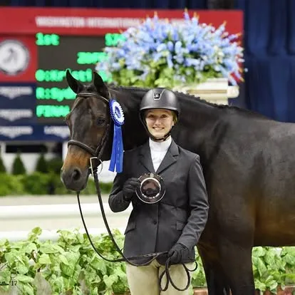 Rider holds trophy beside bay horse with blue ribbon indoors