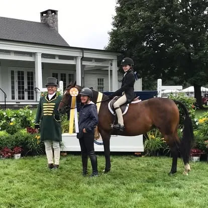 Ringmaster, groom and young rider with bay horse before white house