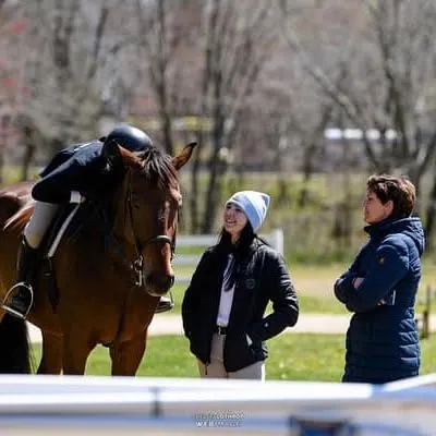 Two women in jackets talk beside a saddled bay horse outdoors