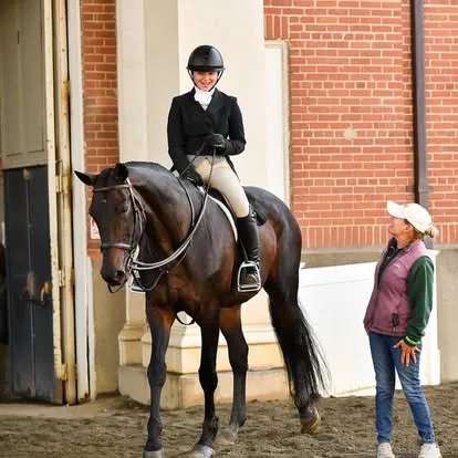 Rider on dark bay horse beside trainer outside brick stable