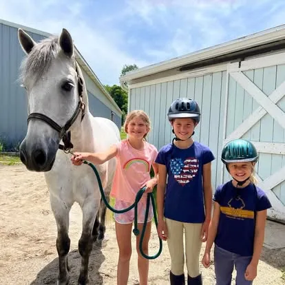 Three helmeted young students holding a grey pony outside a barn
